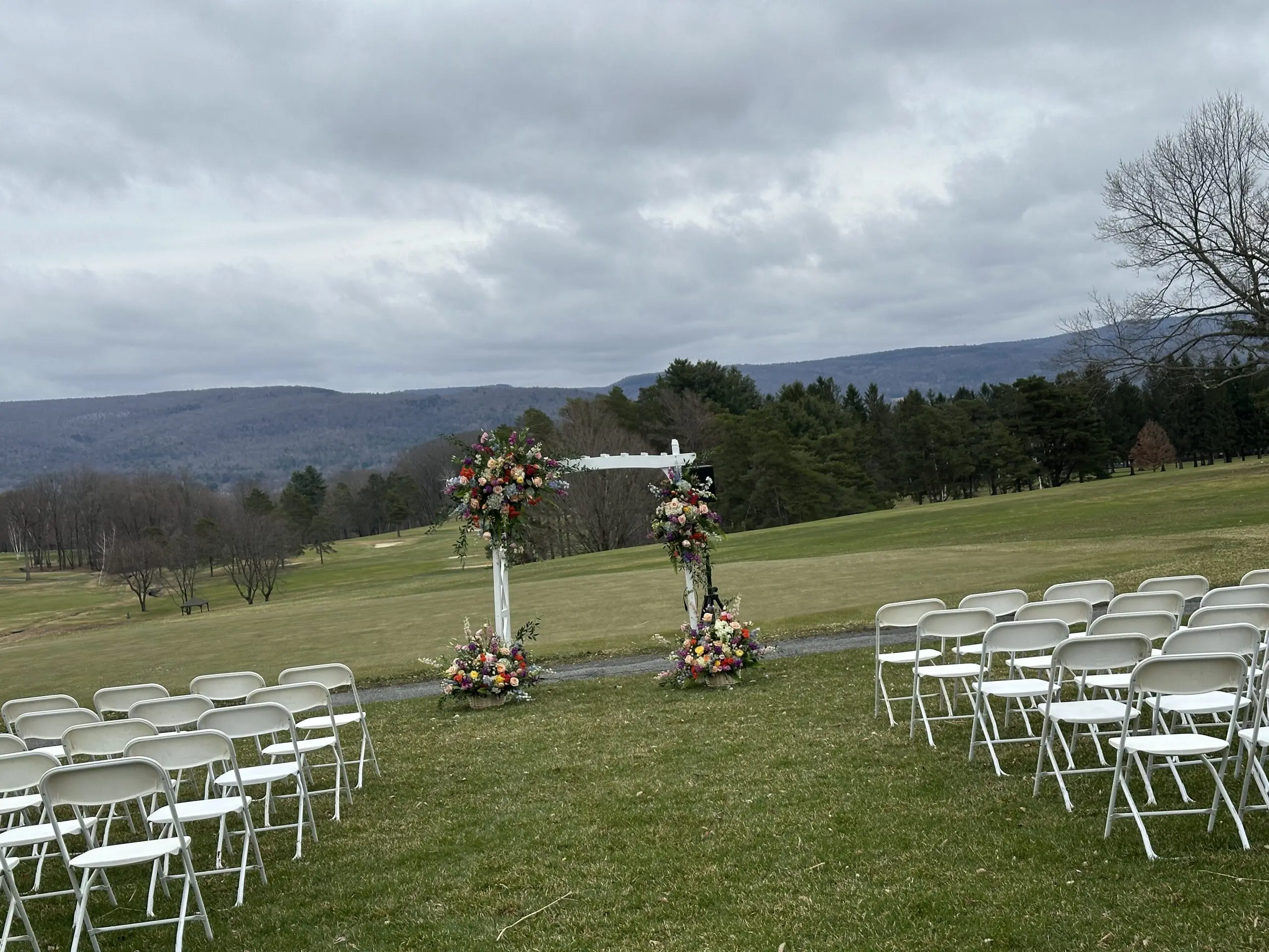 Outdoor wedding setup with decorated arch and white chairs on grassy field.